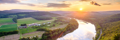 Sun rises over a calm river flowing through scenic valley. Farm fields and forests are green and healthy.